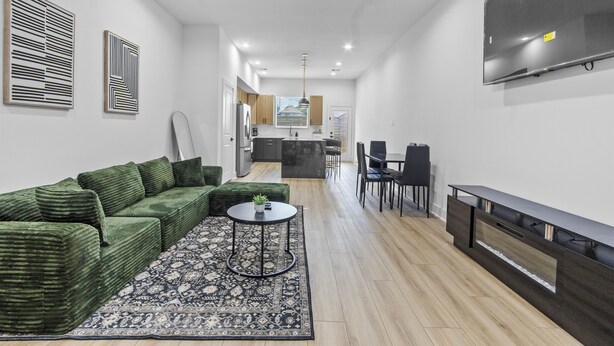 Ein modernes Wohnzimmer mit einem grünen Sofa, einem gemusterten Teppich, einem Fernseher und einem Essbereich mit Stühlen.