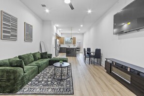 A modern living room with a green sofa, a patterned rug, a TV, and a dining area with chairs. - Firepit BBQ Grill Queen bds sleeps 11 near DWTN with parking (Houston)