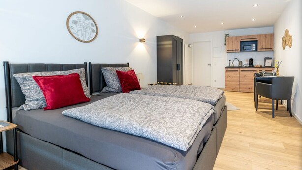 Quarto moderno com cama grande, escrivaninha e cozinha compacta ao fundo.