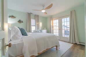 Ein Schlafzimmer mit Bett, Deckenventilator, Fenster mit Vorhängen und Blick ins Freie.