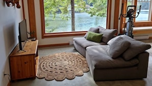 A living room with a large window, a grey sofa, a wooden side table, and a stationary exercise bike. - Charming waterfront Salt Spring Island B&B (Saltspring Island)