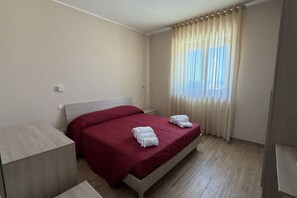 Hotellihuone, jossa on sänky, yöpöytä, ikkuna verhoilla ja vaaleanvärinen lattia.