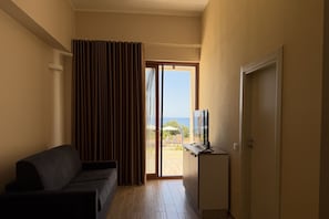 A room with a sofa, a television on a stand, a sliding glass door leading to an outdoor area with a view of the sea.