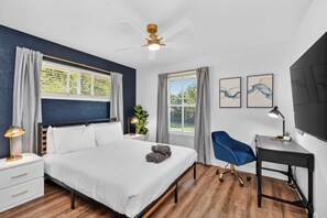 A hotel room with a bed, desk, chair, ceiling fan, window with curtains, and artwork on the wall. - Timeless Charm w/ Pool, Backyard, Patio (5BD/2BA) (Dallas)