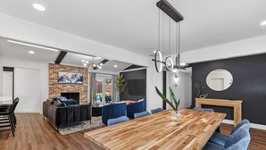 Uma sala de estar moderna com lareira, área de jantar com mesa de madeira e cadeiras azuis, e cozinha visível ao fundo.