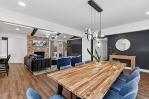 Ein modernes Wohnzimmer mit Kamin, eine Essecke mit Holztisch und blauen Stühlen, und eine im Hintergrund sichtbare Küche.
