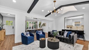 Uma sala de estar moderna com sofá, poltronas, mesa de centro e área de jantar, com cozinha ao fundo.