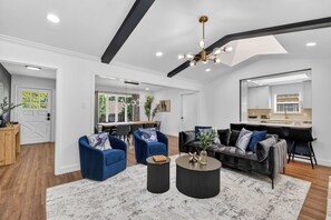 Ein modernes Wohnzimmer mit einer Couch, Sesseln, einem Couchtisch und einem Essbereich, im Hintergrund eine Küche.