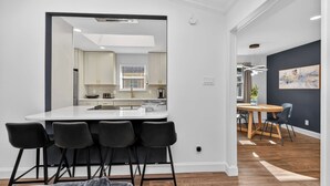 Uma cozinha moderna com armários brancos, uma ilha central com banquetas e um espaço de refeições com parede escura e arte abstrata.