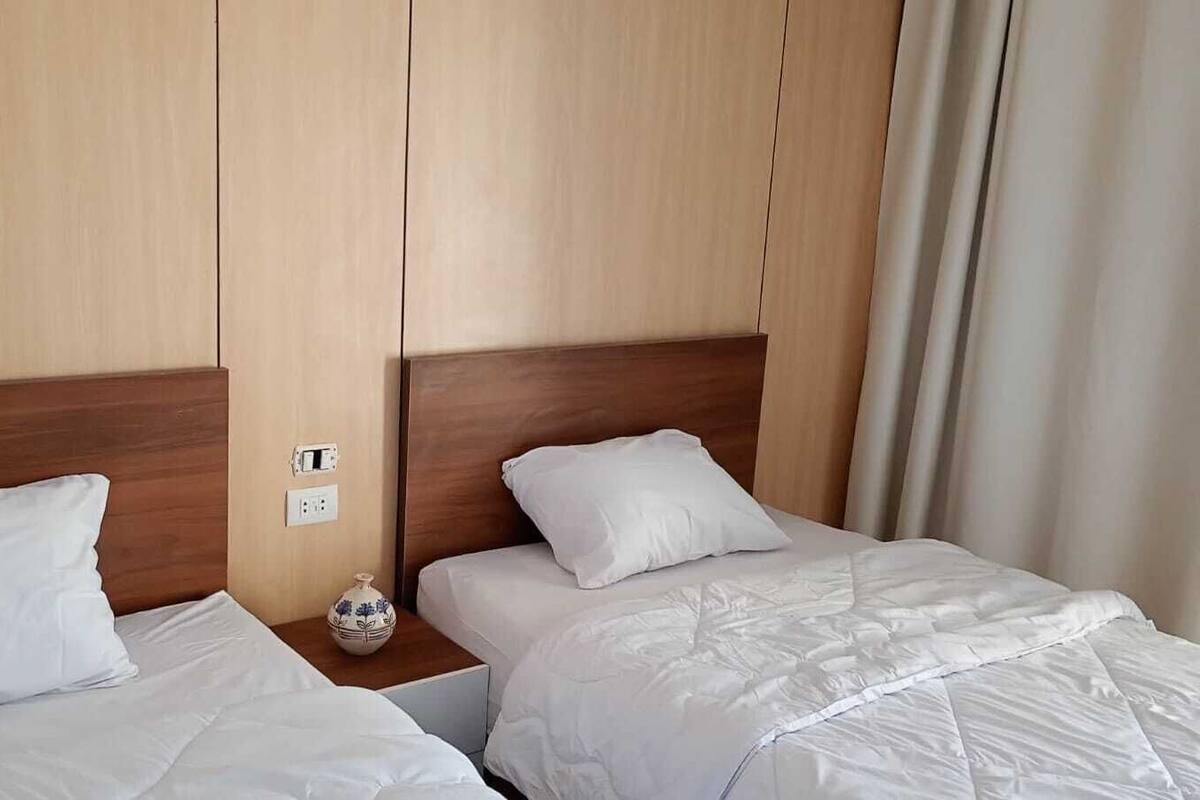 Hotellihuone, jossa on kaksi sänkyä, puiset päätyosat, valkoiset vuodevaatteet, pieni koristeellinen maljakko ja pistorasia seinässä.