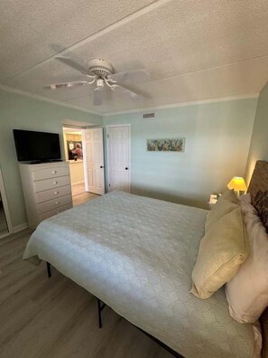 A bedroom with a bed, a ceiling fan, a television, a dresser, and a door leading to another room. - Ocean Front Beach Condo - Windjammer 410 (St. Augustine)