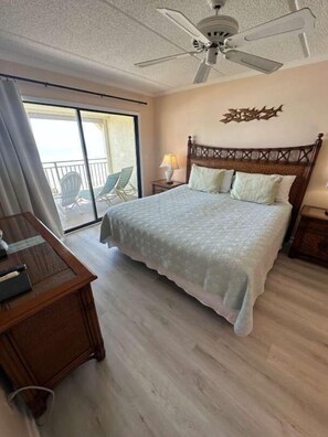A hotel room with a bed, a desk, a ceiling fan, a balcony with chairs, and a decorative wall piece. - Ocean Front Beach Condo - Windjammer 410 (St. Augustine)
