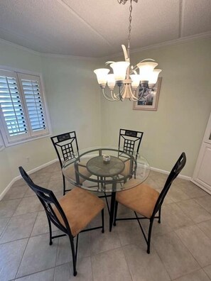 Uma sala de jantar com uma mesa redonda de vidro, quatro cadeiras, um lustre e uma janela com persianas.