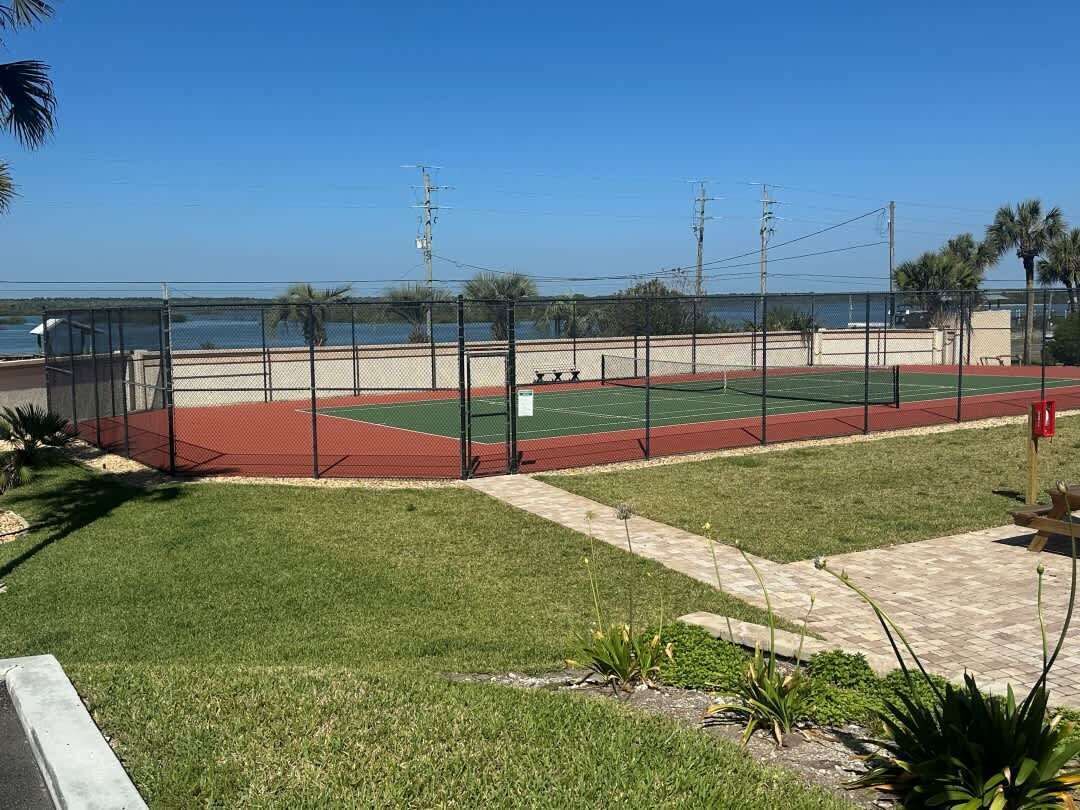 Un court de tennis entouré d’une clôture, avec un chemin et une vue sur l’eau.