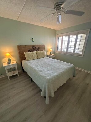 A bedroom with a bed, two bedside tables with lamps, a ceiling fan, and a window with blinds. - Ocean Front Beach Condo - Windjammer 410 (St. Augustine)