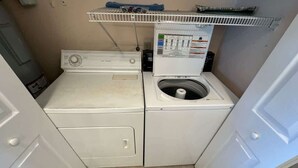 A compact laundry area with a front-loading washer and dryer, a shelf above the dryer, and a tiled floor. - Ocean Front Beach Condo - Windjammer 410 (St. Augustine)