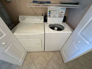 A compact laundry area with a front-loading washer and dryer, a shelf above the dryer, and a tiled floor. - Ocean Front Beach Condo - Windjammer 410 (St. Augustine)