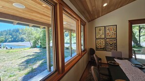 Un espace repas offrant une vue sur un lac et une forêt, avec un plafond en bois et des œuvres d’art botanique encadrées.