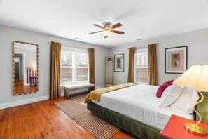 Ein Schlafzimmer mit Bett, Deckenventilator, Fenster mit Vorhängen, Nachttisch mit Lampe und Spiegel an der Wand.