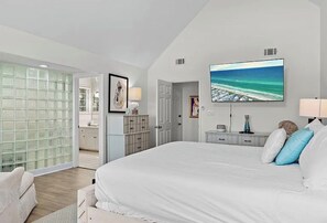 Ein Schlafzimmer mit einem großen Bett, einem an der Wand montierten Fernseher, einem Schreibtisch mit Lampe und Blick auf den Ozean durch das Fenster.
