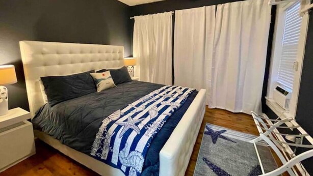 A bedroom with a bed, white curtains, a ceiling fan, and a nightstand with a lamp. - Salty Shark Q1 | Deck Patio (Galveston)