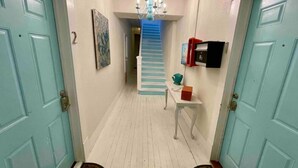 A hallway with light blue doors, a staircase, a chandelier, a small table with a red box, and a wall-mounted device. - Salty Shark Q1 | Deck Patio (Galveston)
