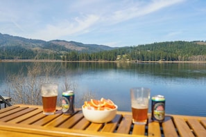 A lakeside view with a wooden deck, a bowl of fruit, and two cans of beer.