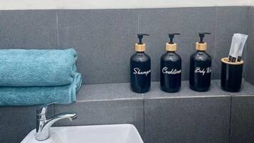 A bathroom with a white sink, a towel, and three black bottles labeled 'Shampoo,' 'Conditioner,' and 'Body Wash' on a shelf.