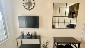 Ett modernt vardagsrum med en platt-TV, ett svart avlastningsbord med växter, ett matbord med stolar, en stor spegel och en klocka på väggen.