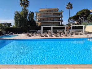 A swimming pool with lounge chairs and a covered area, surrounded by a multi-story building with balconies. - Sea and Sun (Saint-Laurent-du-Var)