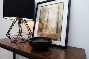 Ein gerahmtes abstraktes Gemälde, eine schwarze Metalllampe und eine dunkle Schale auf einem Holztisch.