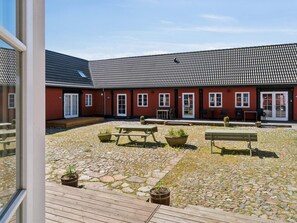 A courtyard with a wooden deck, outdoor seating, and a red building with a tiled roof. - 5 Star Holiday Home in Aakirkeby (Akirkeby)