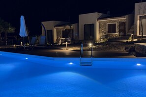 A nighttime swimming pool area with lounge chairs and a covered umbrella, surrounded by modern buildings with stone accents. - Sea-view apartment with a pool (Telegrafo)