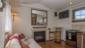 A cozy living room with a fireplace, a sofa, a TV, and a kitchen area. - Carriage House at Roadhouse Lodge: romantic suite, fireplace & lake-view deck (Twin Lakes)