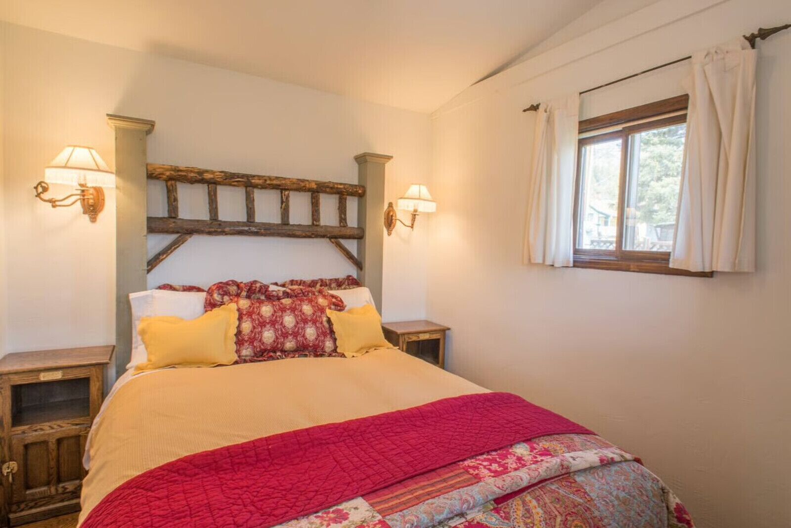 A bed with a red quilt, two bedside tables with lamps, a wooden headboard, and a window with curtains.