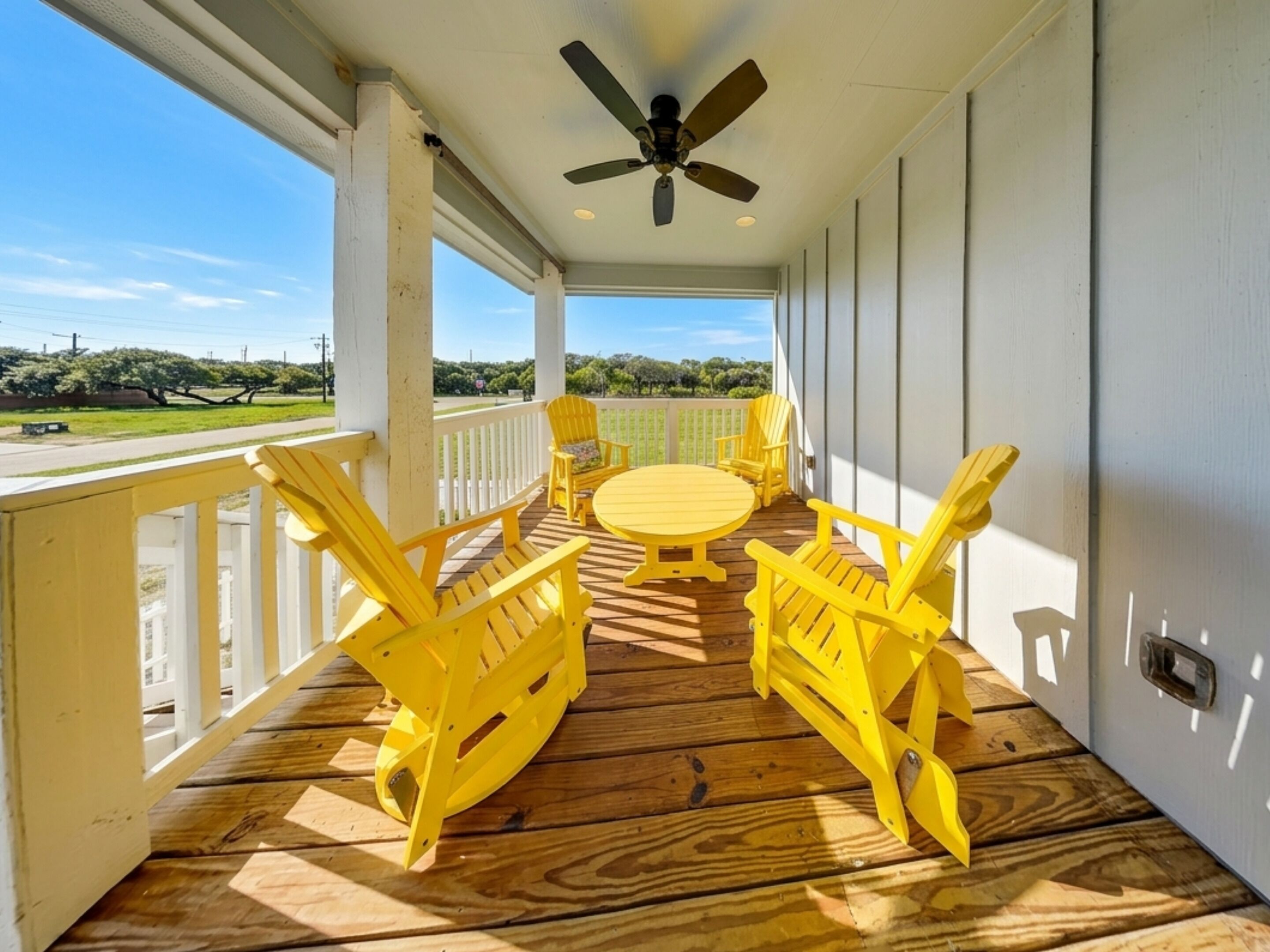A covered outdoor area with a ceiling fan, wooden deck, yellow Adirondack chairs, a round table, and a view of a road and greenery.