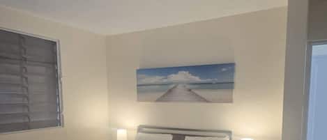 A bedroom with a bed, two bedside tables with lamps, a painting of a beach, and a ceiling light fixture.