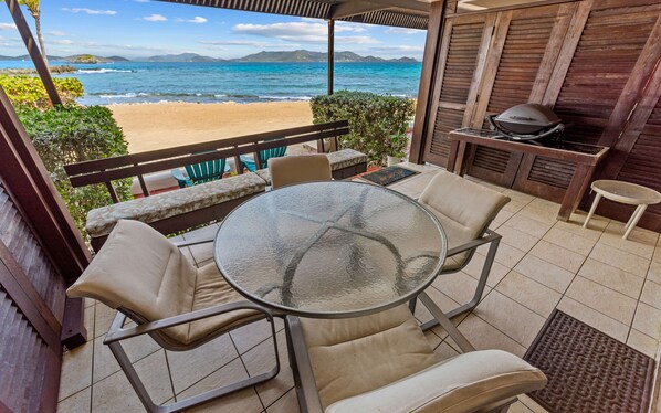 A patio with a glass table and chairs, a barbecue grill, and a view of the beach and ocean.