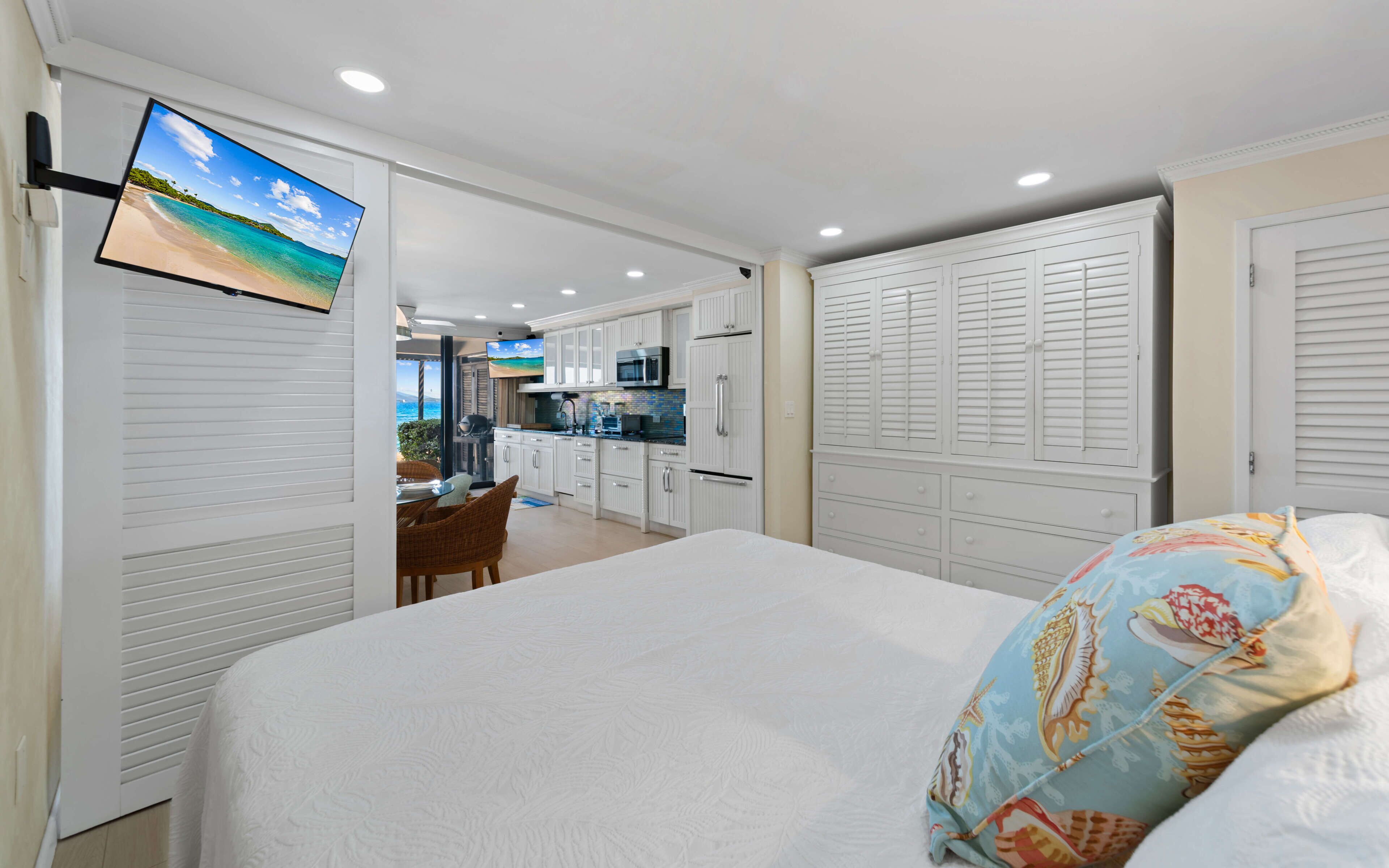 Un dormitorio con cama, televisor montado en una puerta corredera, kitchenette y vista a la playa.