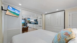 Quarto com cama, TV fixada numa porta deslizante, cozinha compacta e vista para a praia.