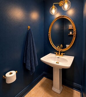 A bathroom with a white pedestal sink, gold fixtures, a round mirror with a gold frame, a towel hanging on a towel rack, and a toilet paper holder.