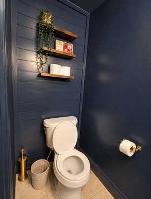 A bathroom with a toilet, a trash can, a toilet brush, and a shelf with a plant and toilet paper.