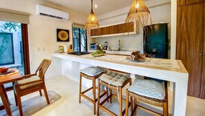 Una cocina con una isla central, taburetes de bar, lámparas colgantes y una zona de comedor con mesa y sillas.