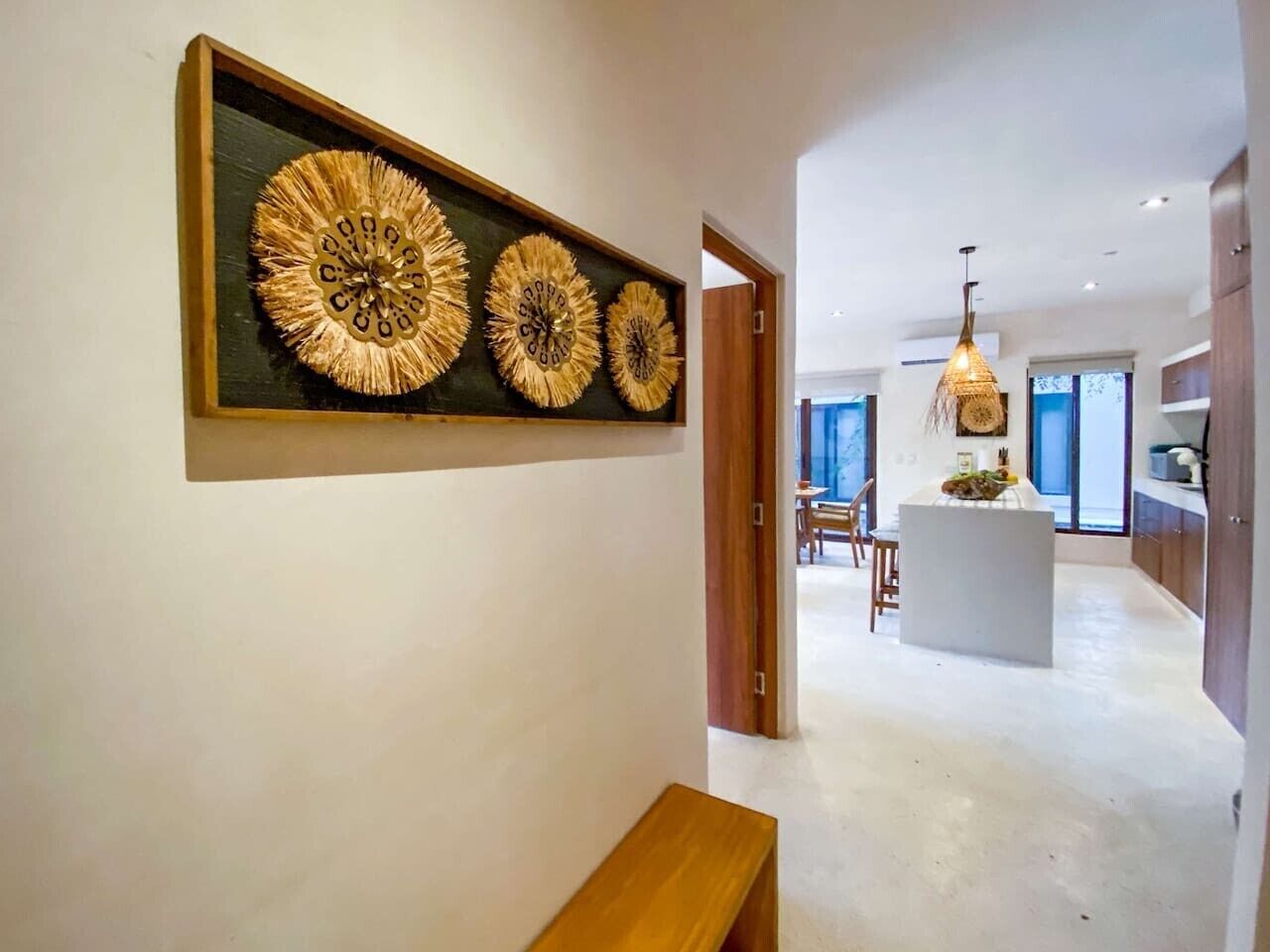 Un pasillo con un banco de madera, un reloj en la pared, y una zona de cocina con una isla central e iluminación colgante.