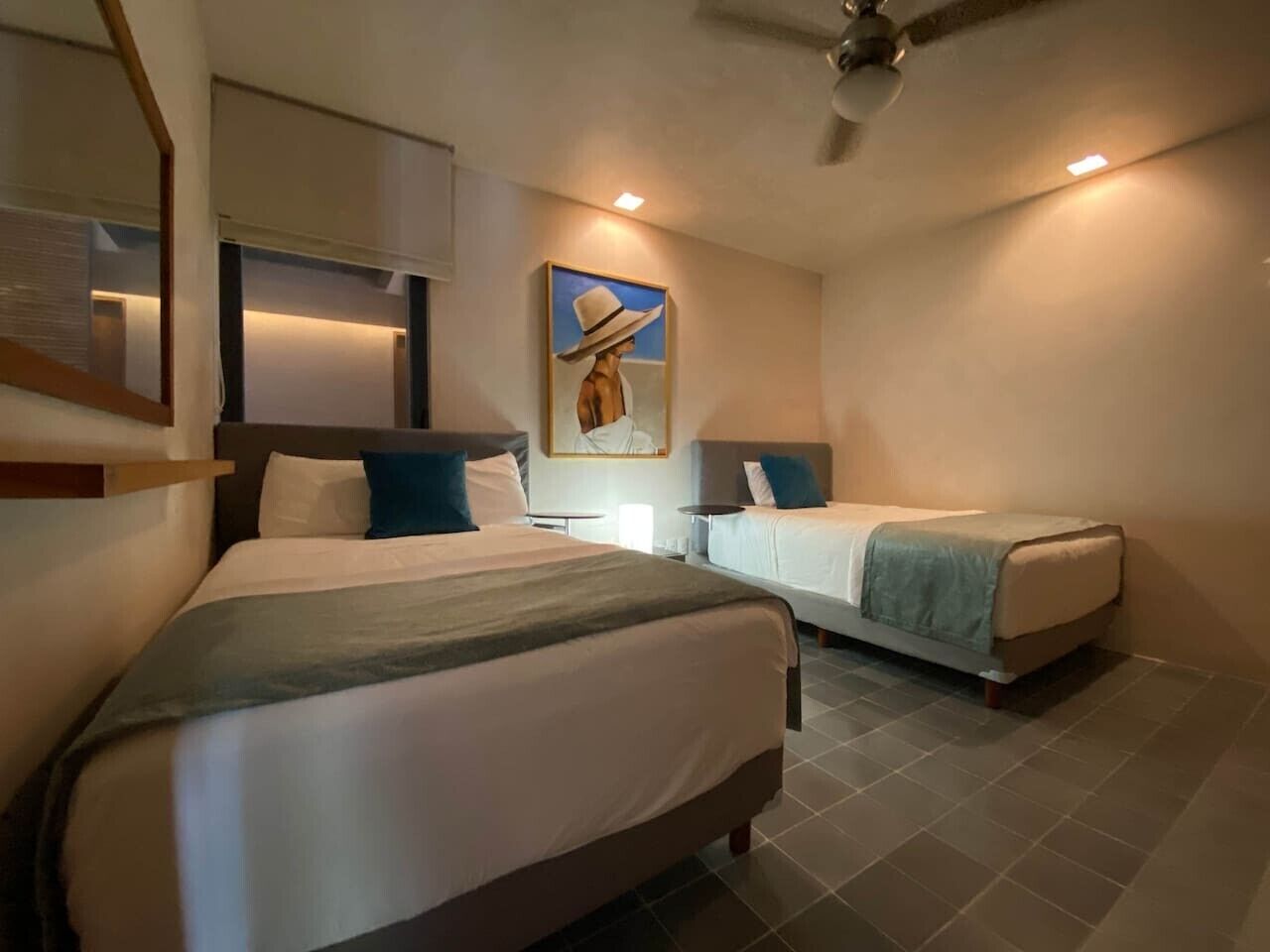 A hotel room with two beds, a ceiling fan, a painting on the wall, and a small table.
