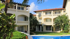 A resort with a swimming pool, palm trees, and a two-story building with balconies. - Ground floor Condo, Steps from Coco Beach & 5th Av (Playa del Carmen)