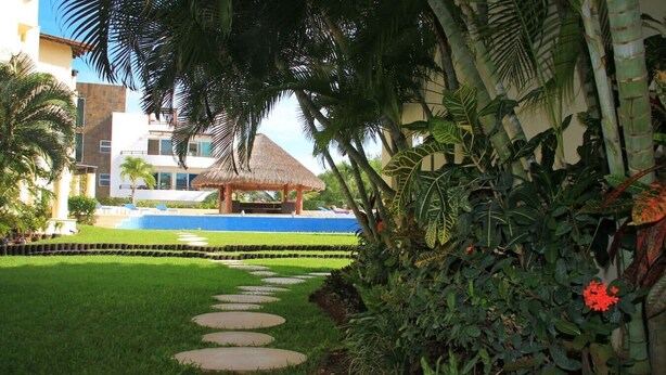 A pathway leading to a thatched-roof structure surrounded by lush greenery and a pool. - Ground floor Condo, Steps from Coco Beach & 5th Av (Playa del Carmen)