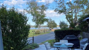 A patio with outdoor furniture, a grill, and a view of a road and trees. - Cozy 2-bedroom waterfront cottage with central AC, patio, BBQ, Private beach (Hampton Bays)