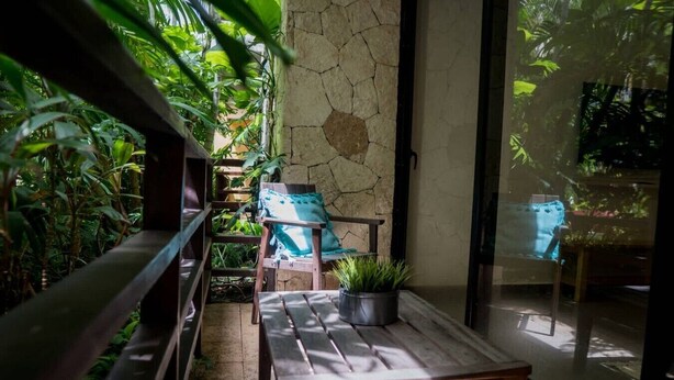 A sunlit patio with wooden furniture, a stone wall, and lush greenery. - Lunada: Bright Stay with Rooftop Amenities (Playa del Carmen)