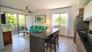 Et moderne køkken med en central ø, et spiseområde med en grøn sofa og en balkon med udsigt til grønne områder.